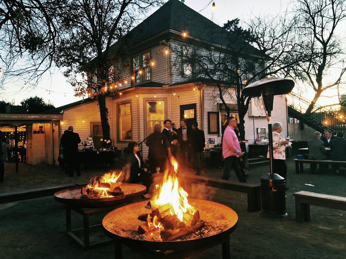 People socialize around fire pits in a lit gravel yard.