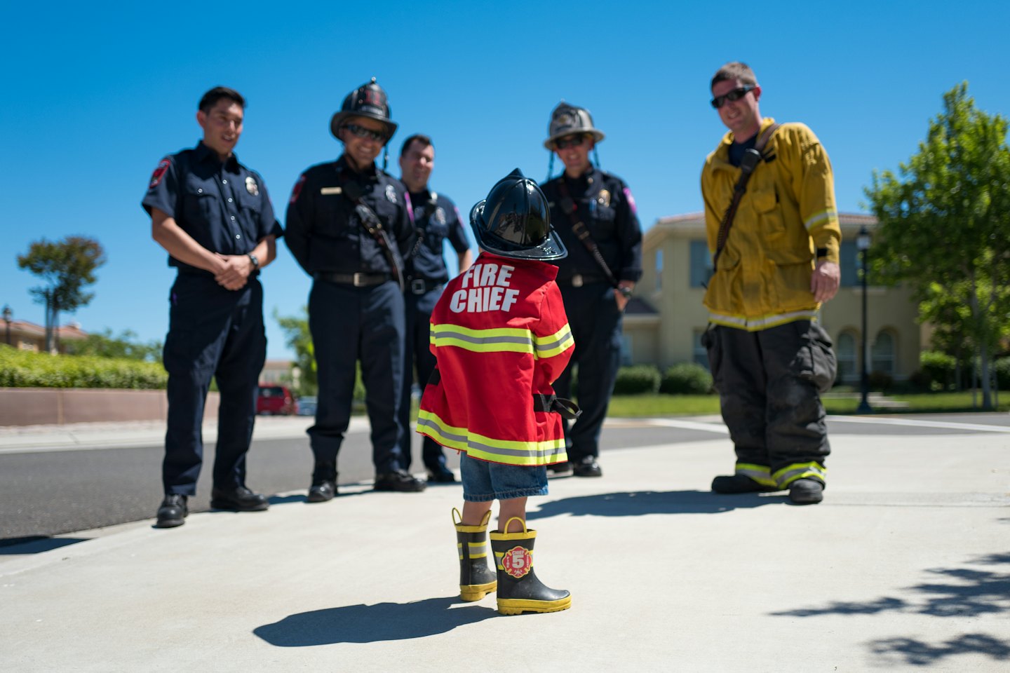 Child wearing a firefighter uniform stands before a group of firefighters.